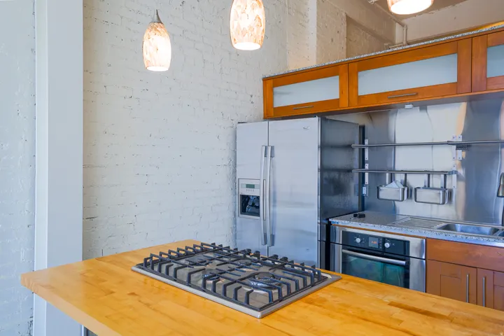 Exposed brick wall, stainless steel refrigerator, built-in gas cooktop, wood countertops, and modern pendant lighting