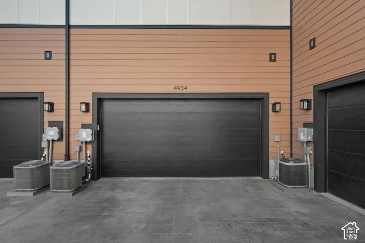 Tan horizontal siding with black trim, multiple black garage doors, gray stone-finish pavement, and exterior wall sconces