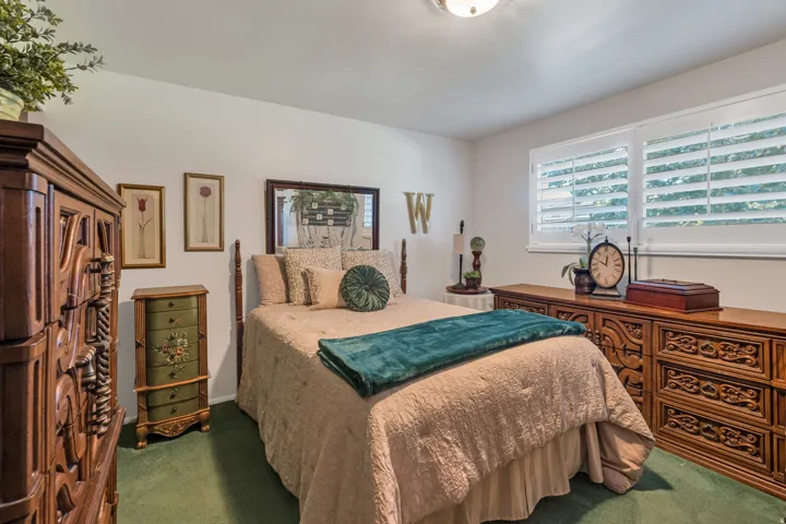 Recessed lighting illuminates the room, featuring a window with plantation shutters and green carpet flooring