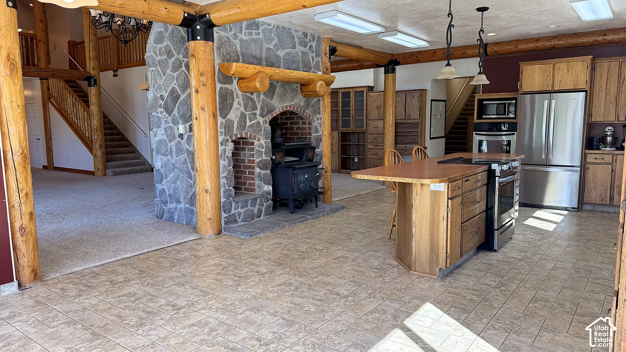 Stone fireplace with a log mantel and wood-burning stove, exposed log structural columns, wood cabinetry, tile flooring, and stainless steel appliances