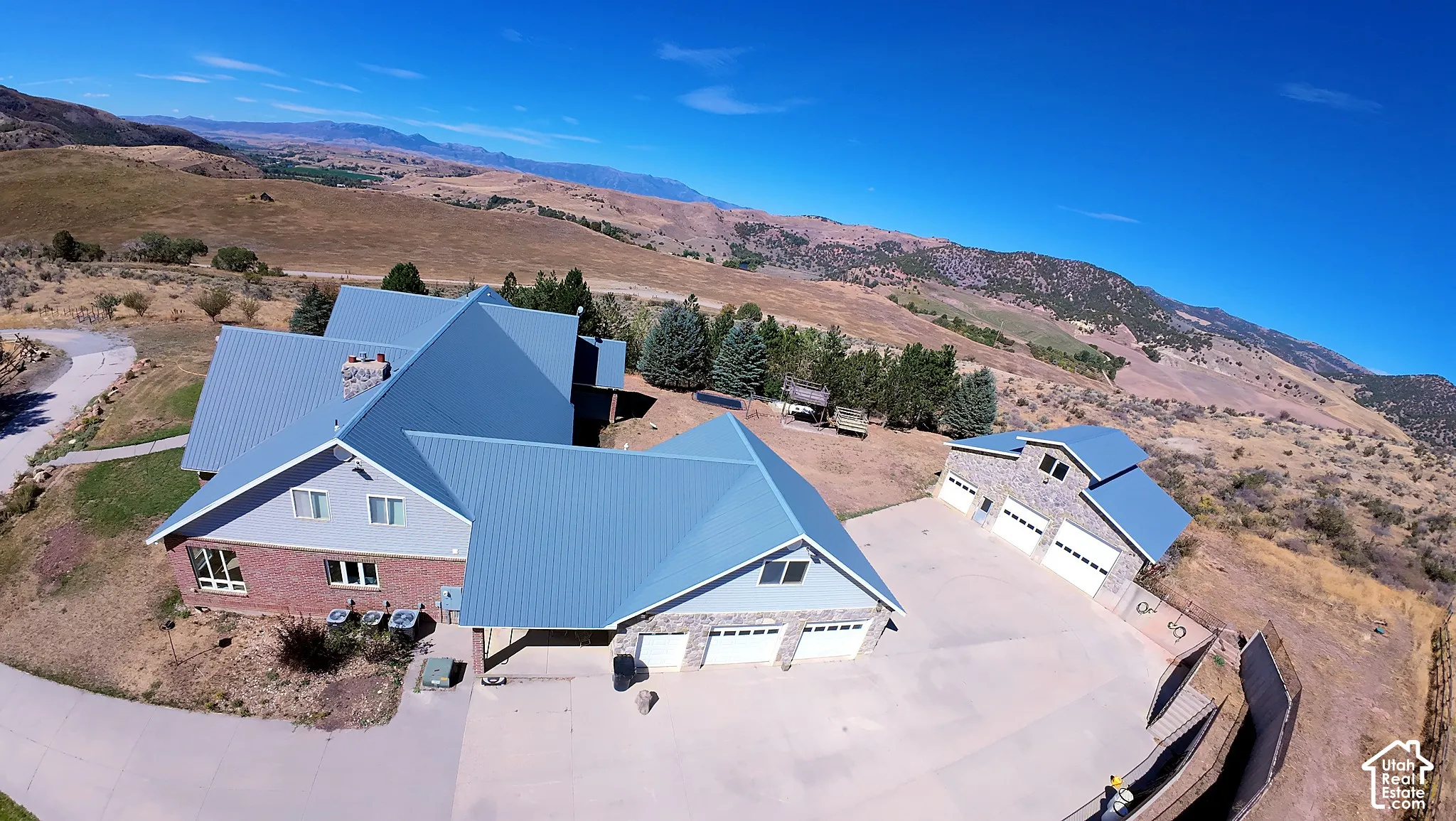 Aerial perspective of a multi-structure property featuring a residence with a metal roof, brick and siding exterior, an integrated three-car garage, and a detached secondary garage