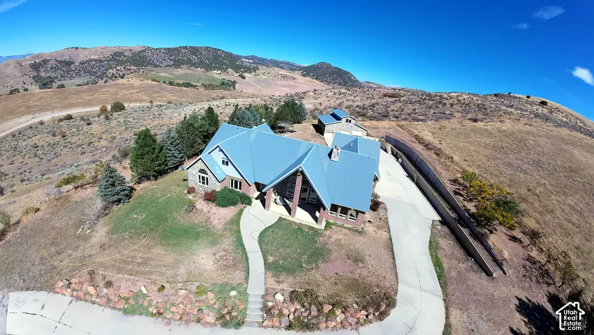 Aerial perspective of a multi-gabled residence featuring a metal roof, stone masonry siding, an expansive concrete driveway, a secondary detached structure, and mountainous terrain