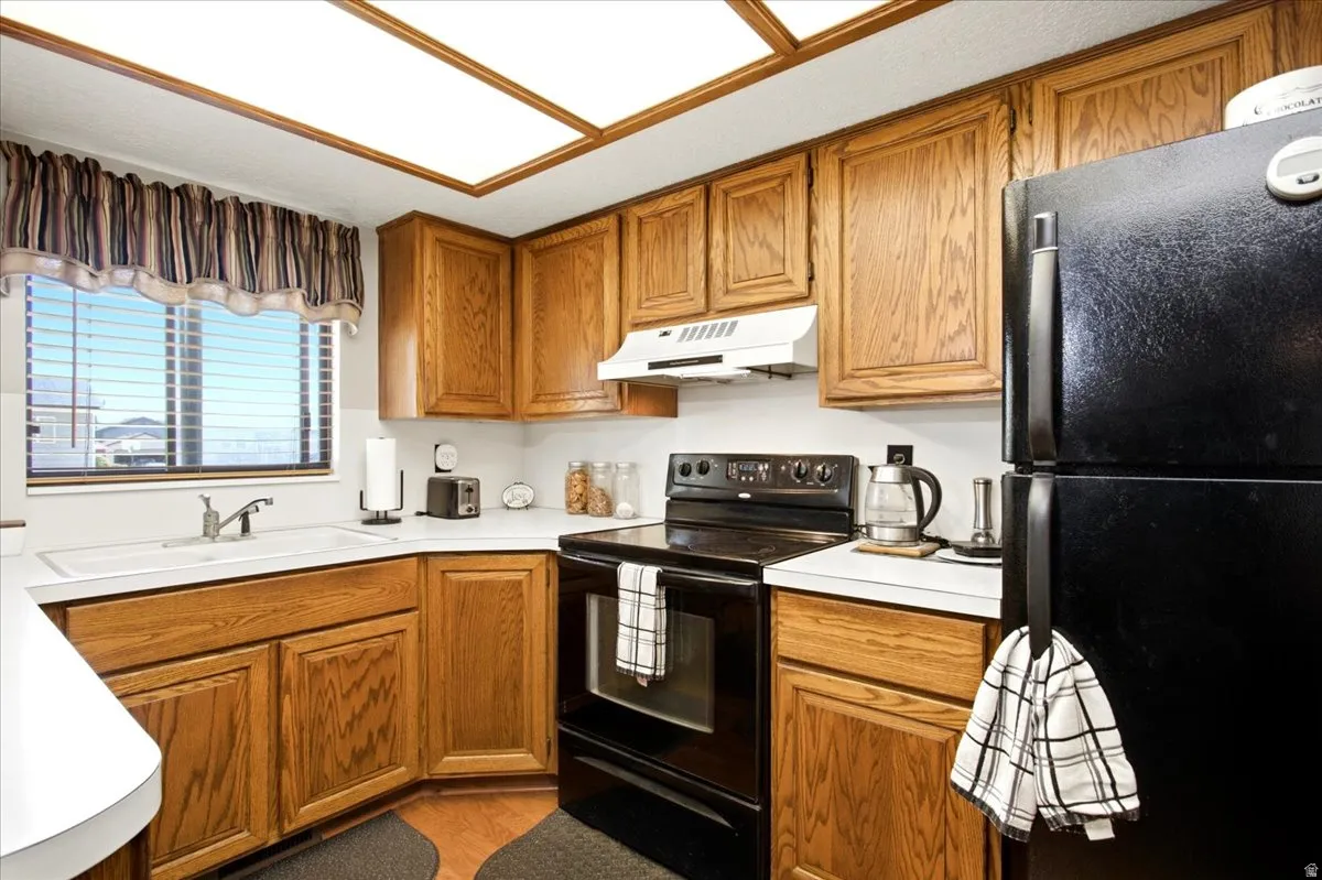 Kitchen featuring wood cabinetry, white countertops, a corner sink with a window, and black appliances including a refrigerator and range