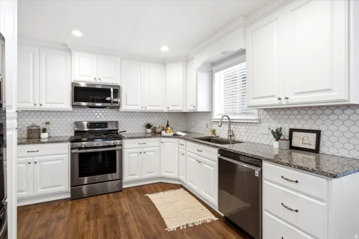 White cabinetry, stainless steel appliances, patterned tile backsplash, dark speckled countertops, and wood-finish flooring