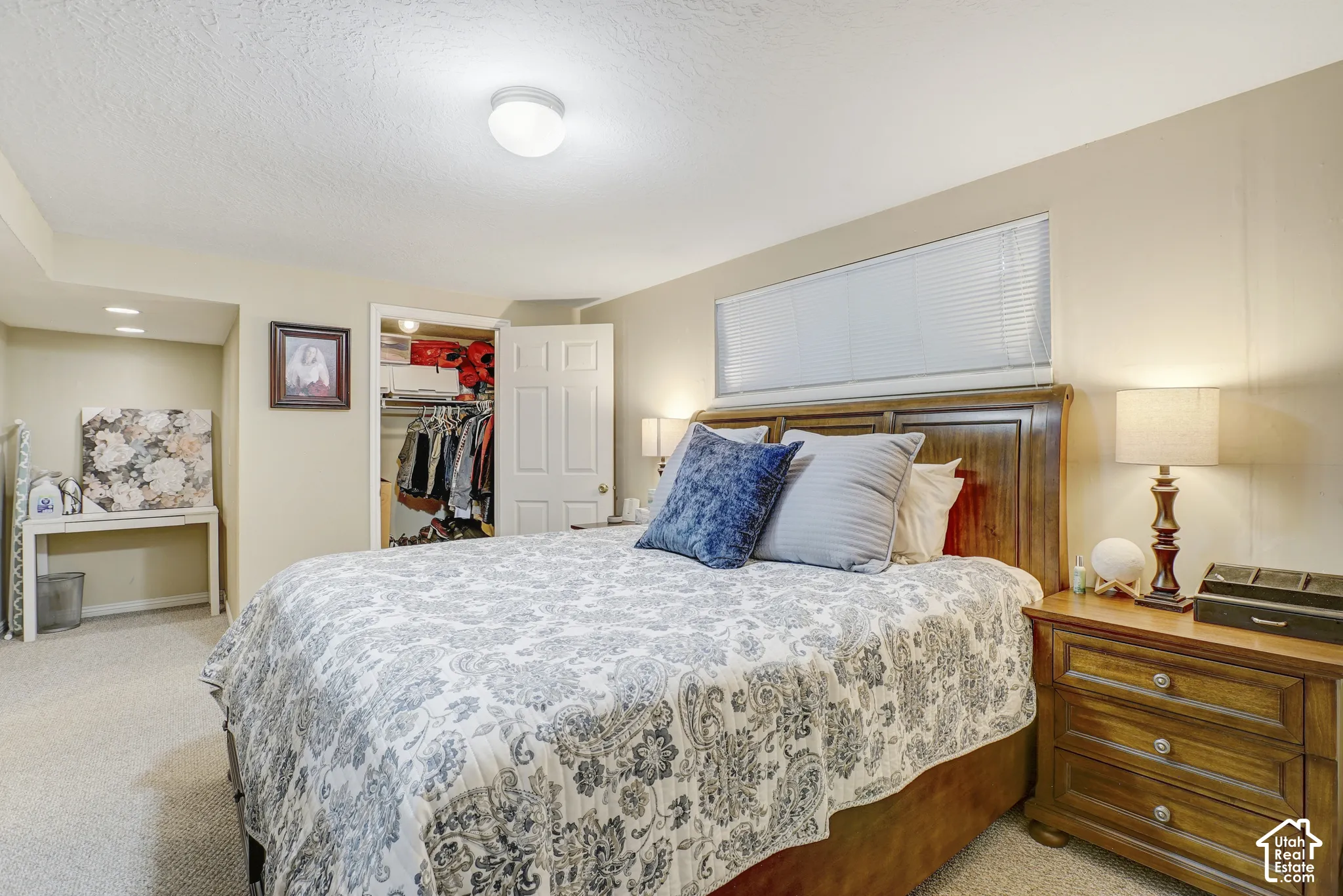 Carpeted flooring with a wood headboard, wood-finish nightstand, and a horizontal window with blinds