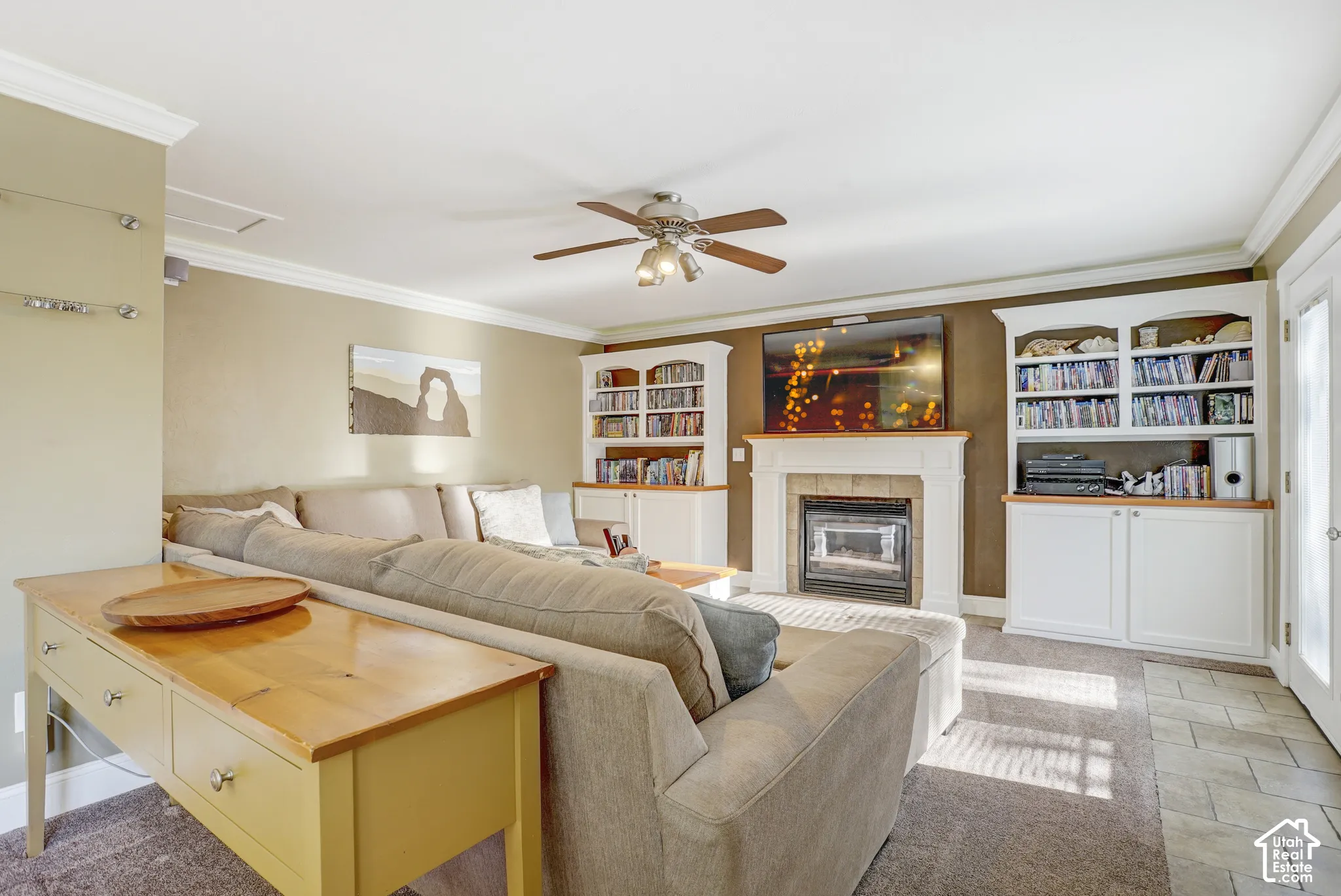 Living area featuring a fireplace with a mantel, built-in shelving, a ceiling fan, and crown molding