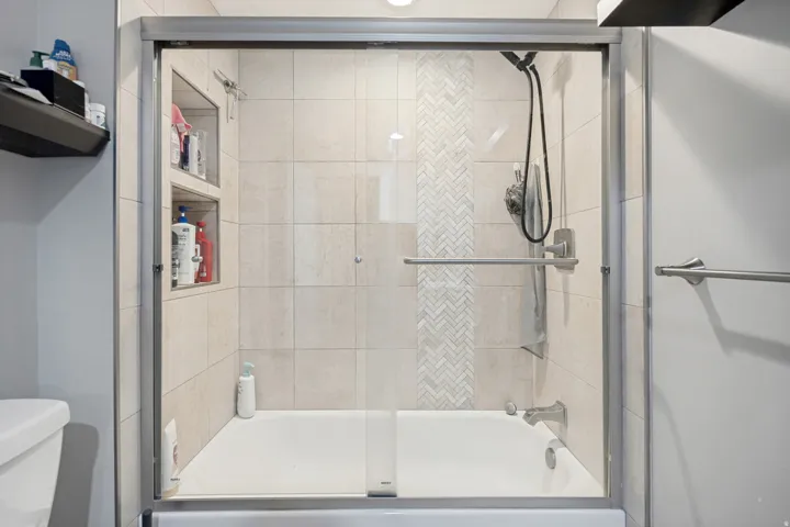 Bathtub with clear glass sliding doors, tiled surround with decorative herringbone accent, built-in shower niches, and brushed nickel fixtures