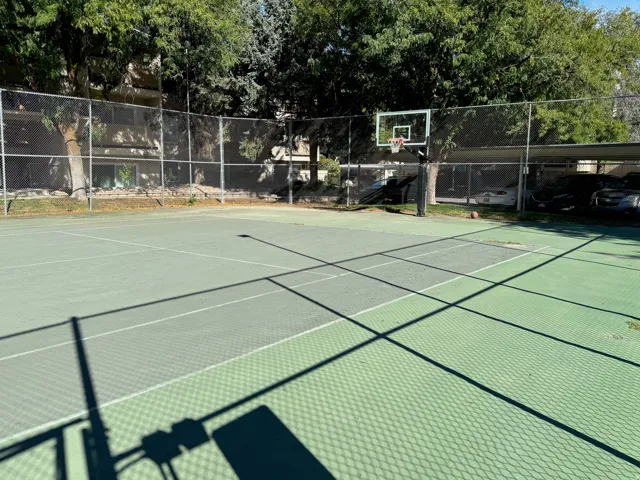 Multi-sport court featuring a basketball hoop and tennis court lines, surrounded by chain-link fencing and mature trees