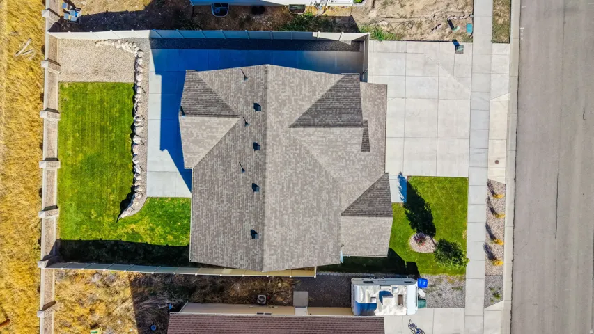 Pitched roof with asphalt shingles, concrete driveway, manicured lawn, and stone retaining wall