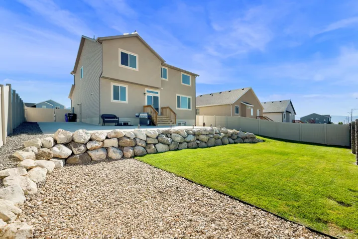 Expansive backyard featuring a manicured lawn, a gravel patio area, a rock retaining wall, and a concrete patio