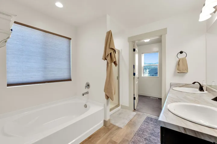 White soaking tub with chrome fixtures, wood-finish flooring, dual vanity with undermount sinks, and dark bronze faucets