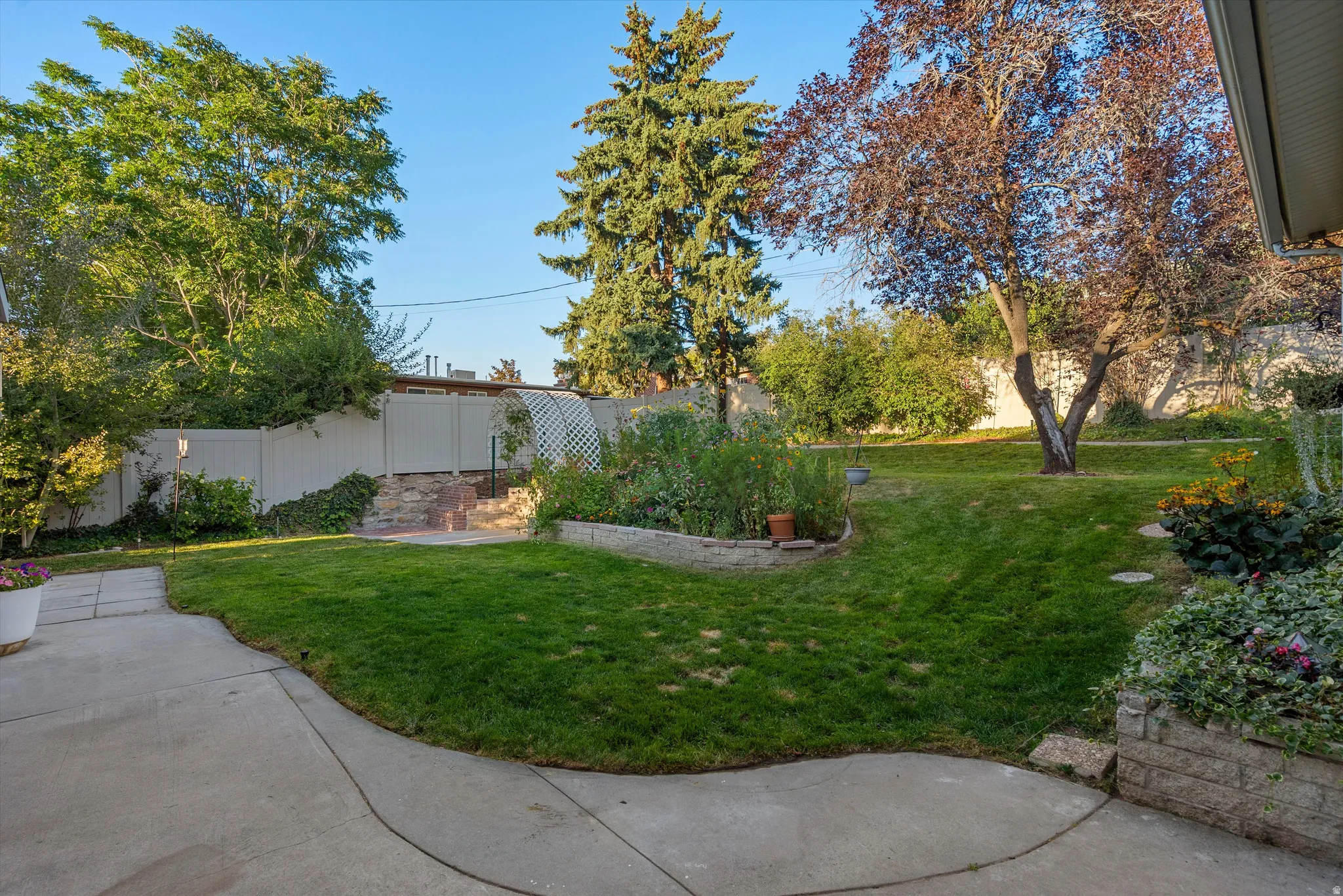 Expansive backyard featuring a concrete patio, lush lawn, mature trees, garden beds, and privacy fencing
