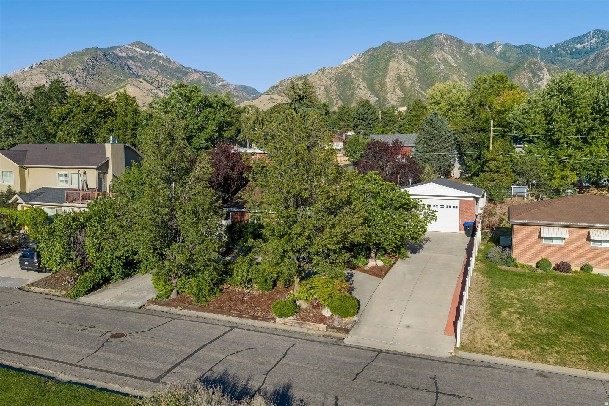 Two-car garage with white door, concrete driveway, front yard with mature trees, and surrounding mountains