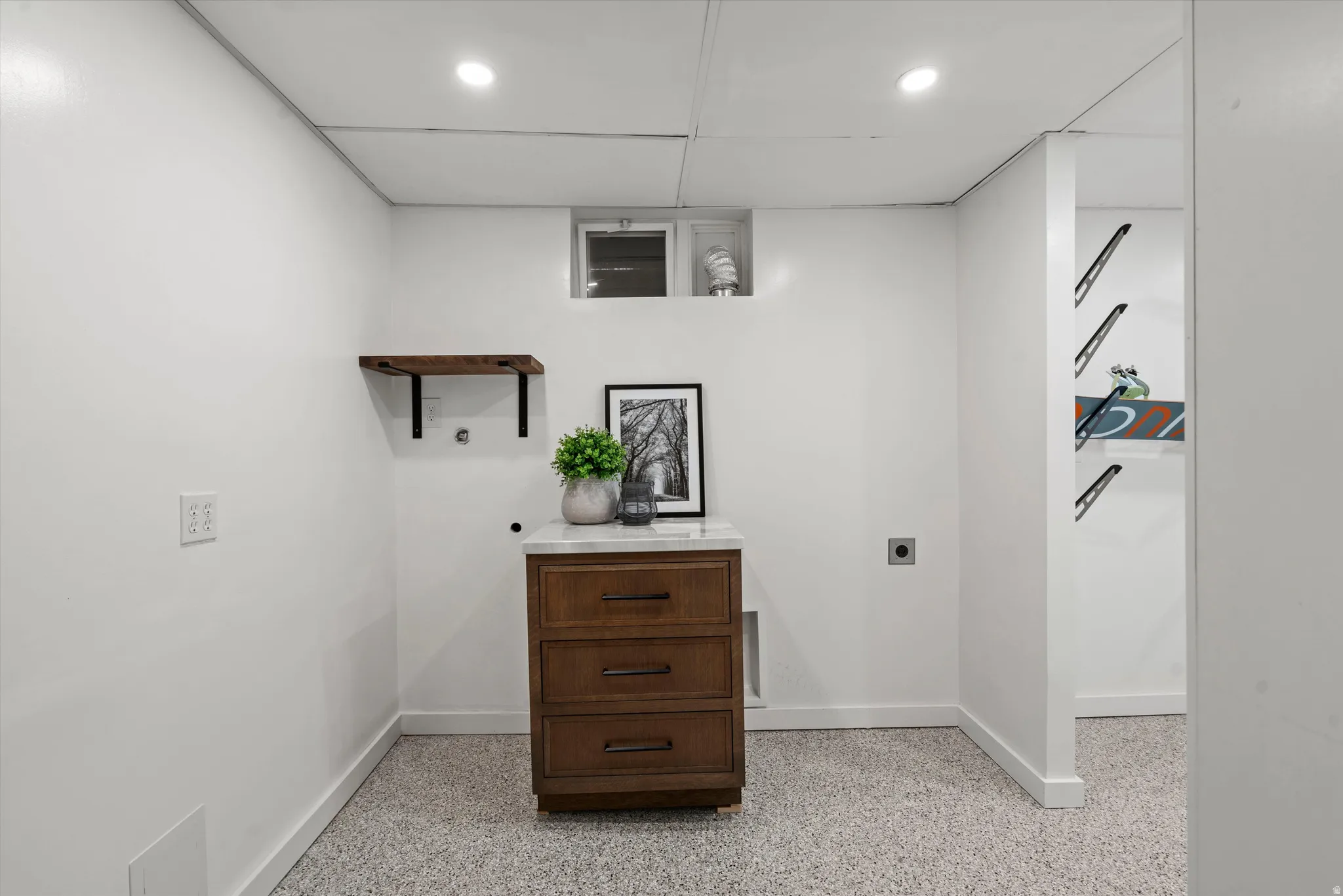Recessed lighting illuminates the space, featuring a floating wood shelf and a built-in wood-finish cabinet with a light-colored countertop epoxy flooring