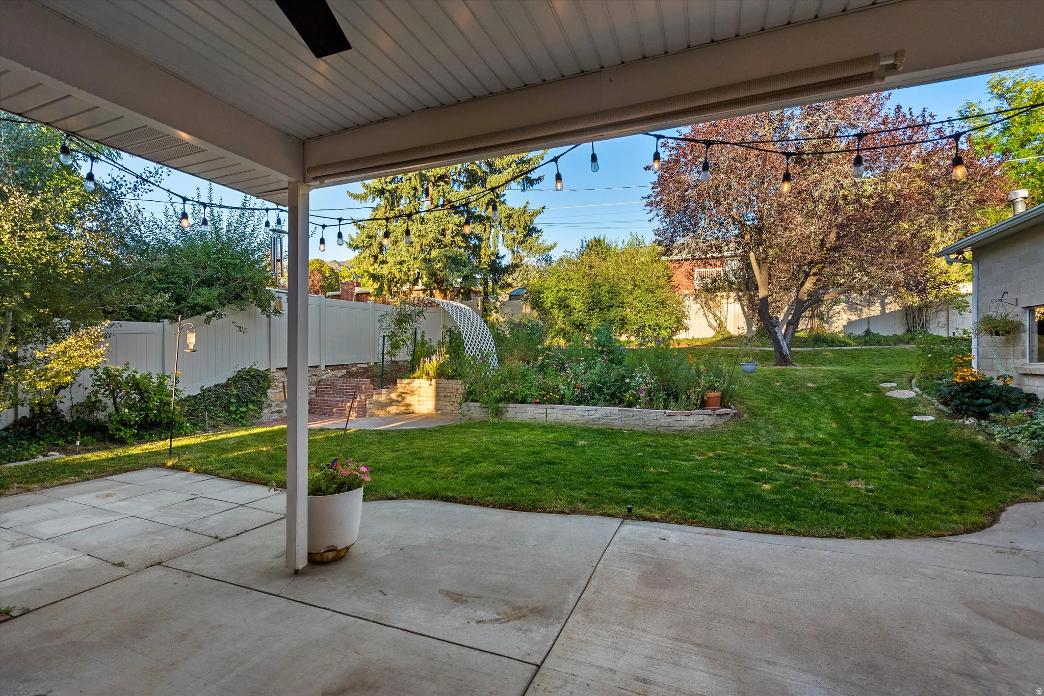 Covered patio with concrete slab, expansive lawn, mature perimeter trees, and raised garden beds
