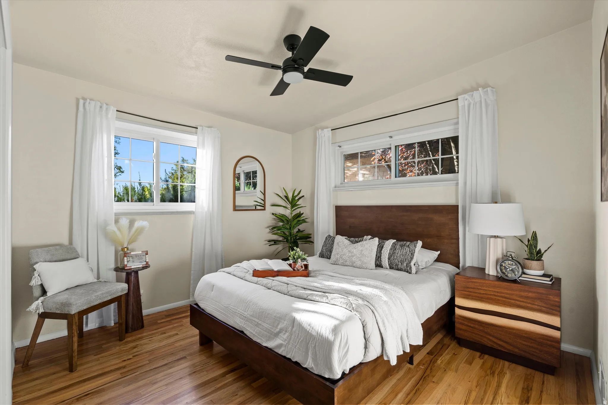 Primary Bedroom #1 Hardwood flooring, two windows with white window treatments, a dark wood headboard, and a black ceiling fan