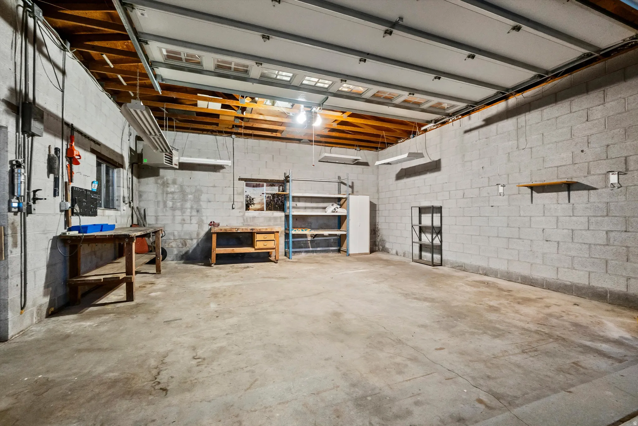 Concrete block wall construction, exposed wooden ceiling beams, concrete flooring, overhead fluorescent lighting, and a roll-up garage door. Garage heater.