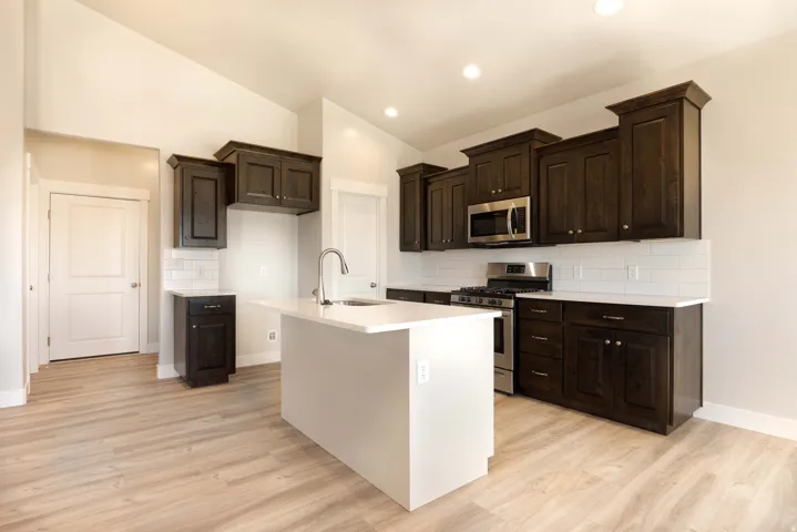 Kitchen featuring dark wood-finish cabinetry, stainless steel appliances, a center island with an undermount sink, white countertops, and a subway tile backsplash