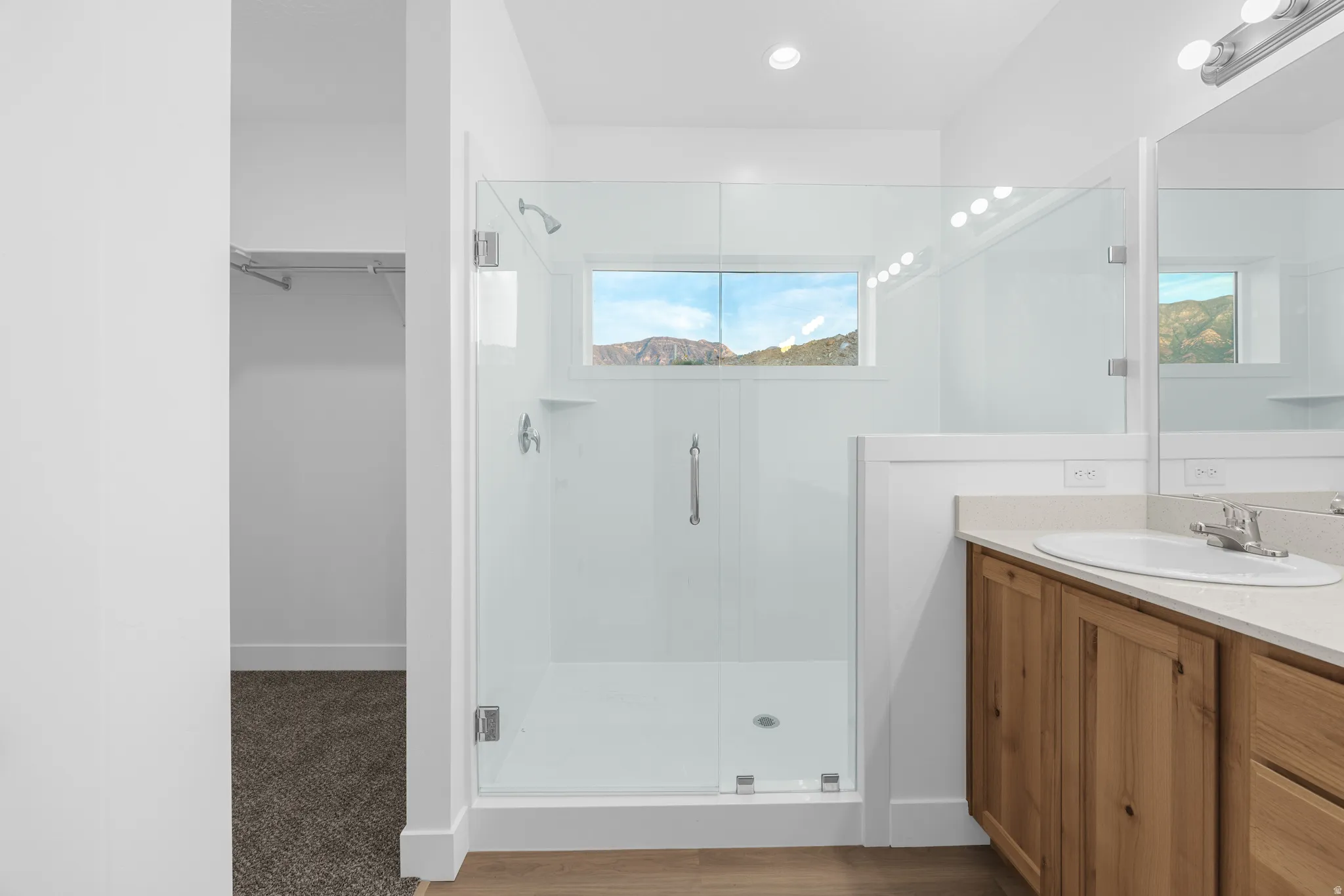 Glass-enclosed shower with a transom window, wood-finish flooring, single vanity with a solid surface countertop, and recessed lighting