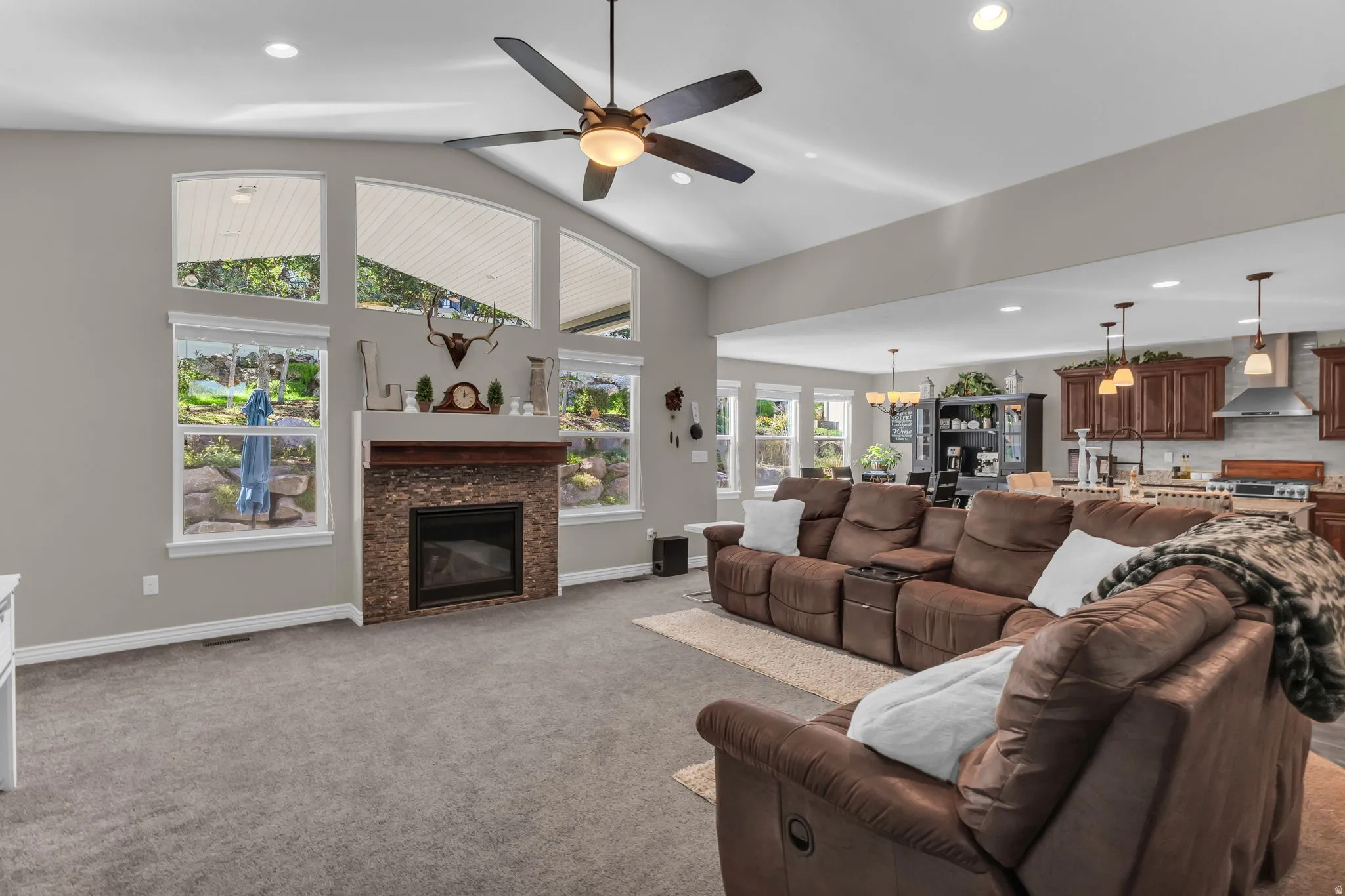 Vaulted ceilings with recessed lighting, stone-clad fireplace, and an open-concept design to the kitchen featuring wood cabinetry and stainless steel appliances