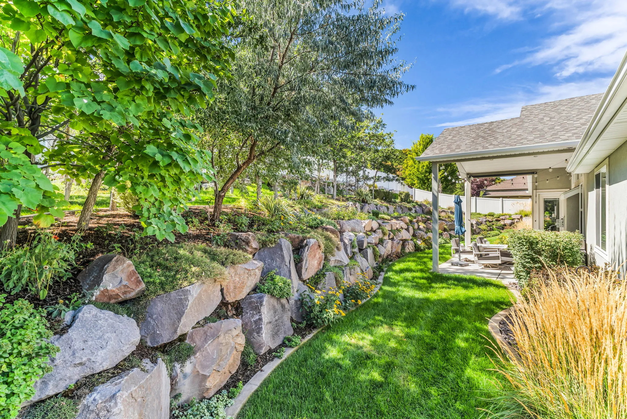 Terraced landscaping with a stacked stone retaining wall, mature trees, manicured lawn, and covered outdoor patio