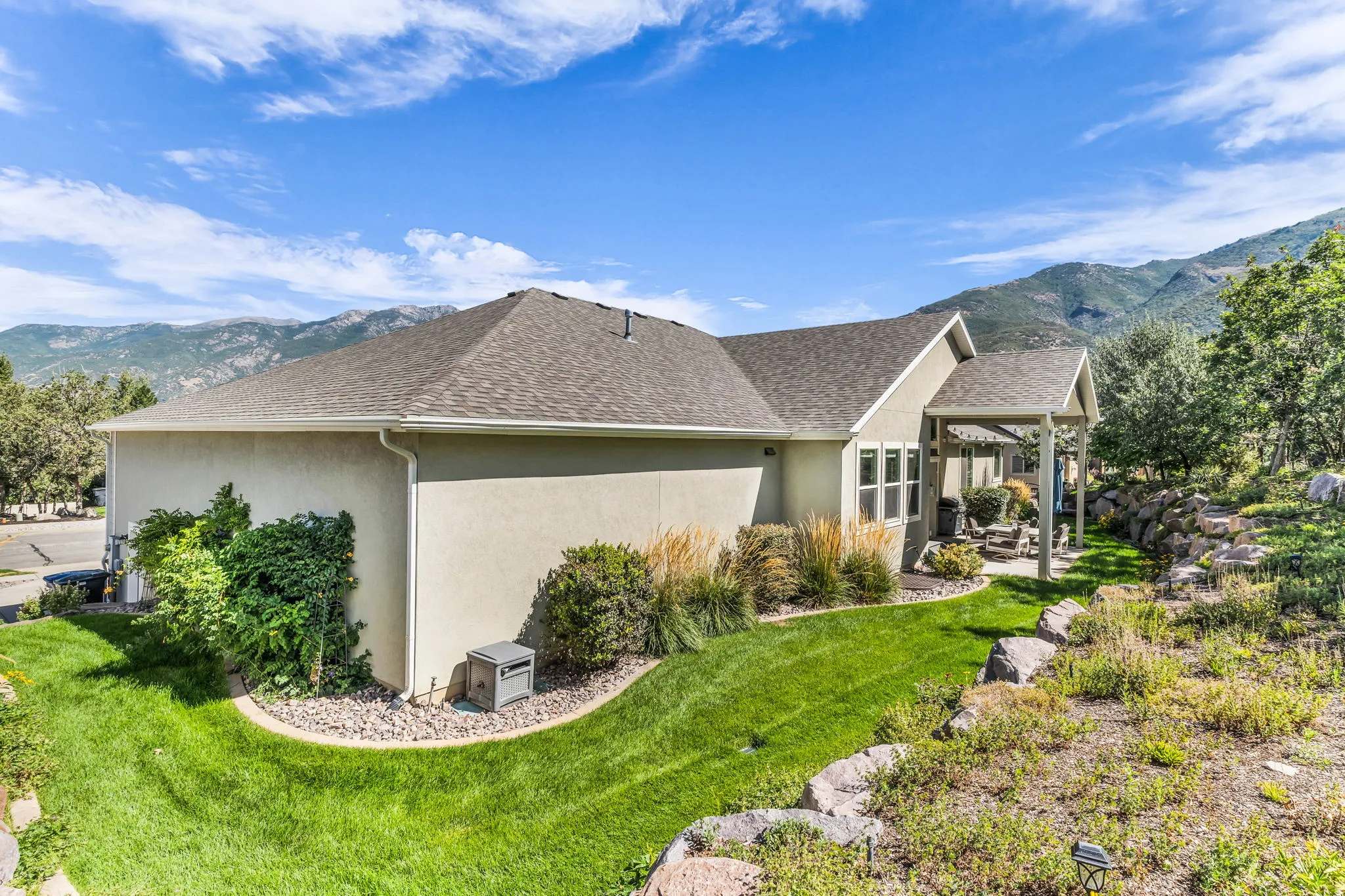 Stucco exterior with a pitched shingle roof, featuring a covered patio, terraced landscaping, and a manicured lawn