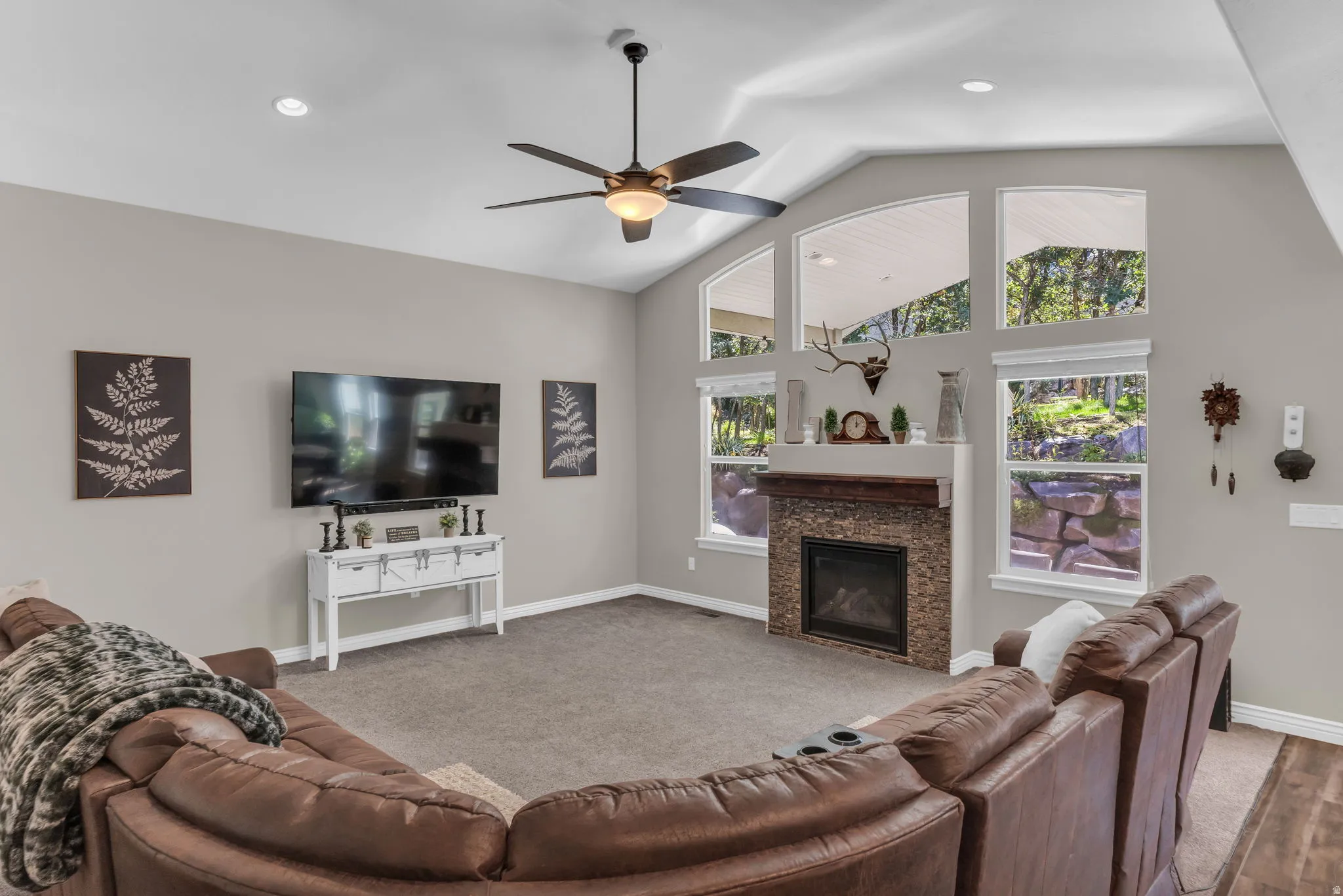 Vaulted ceiling with recessed lighting and ceiling fan, stone-surround fireplace with wood mantel, and expansive windows