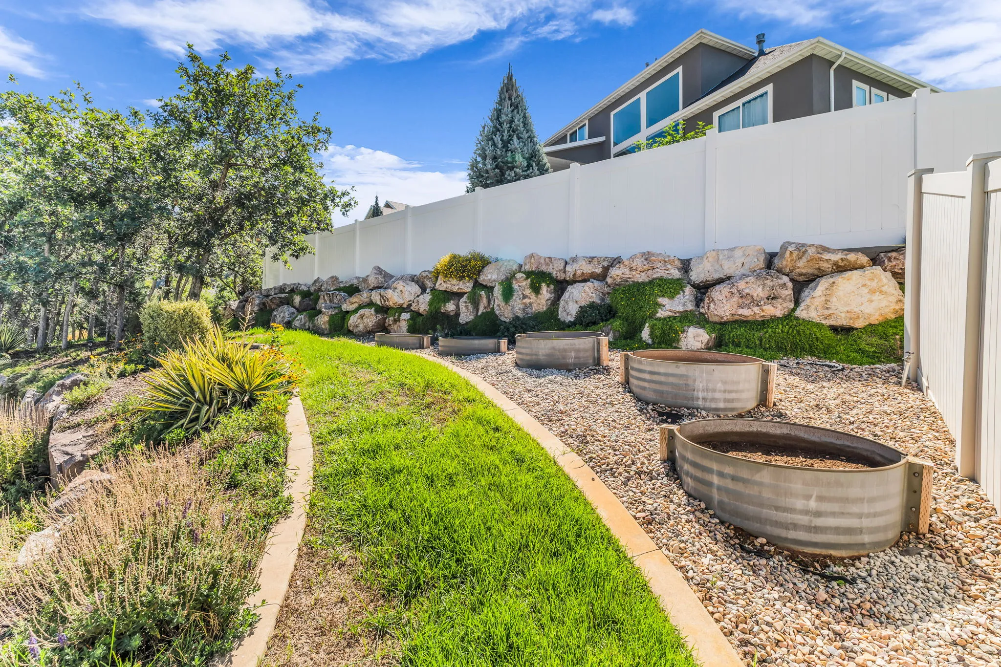 Terraced landscaping with a rock retaining wall, mature trees, and a white privacy fence