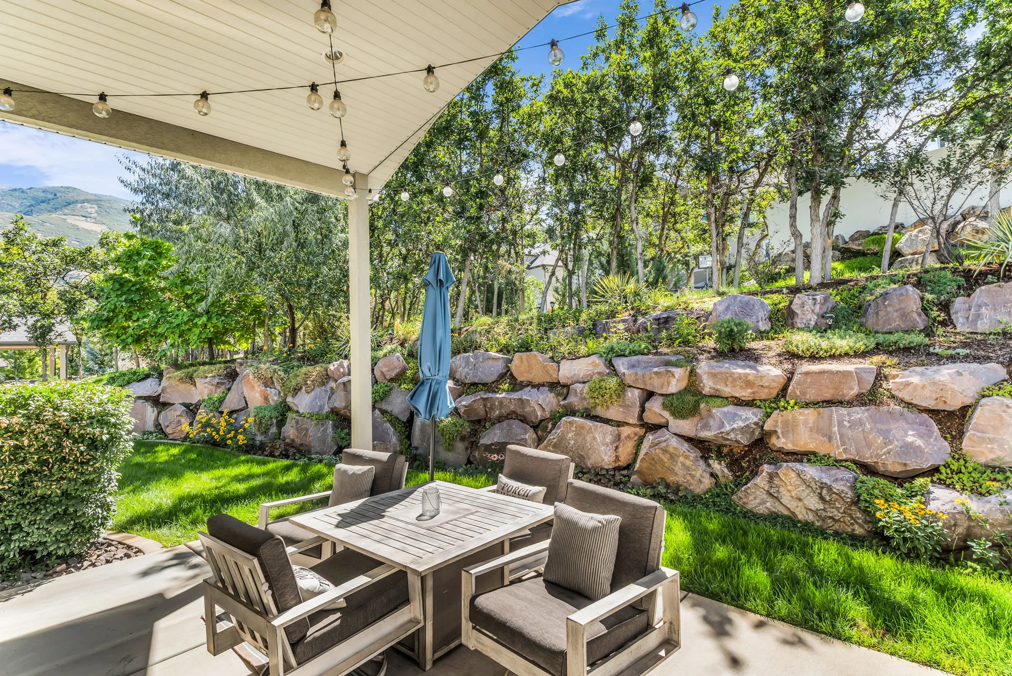 Covered patio with integrated string lighting, expansive rock retaining wall, mature trees, and manicured lawn