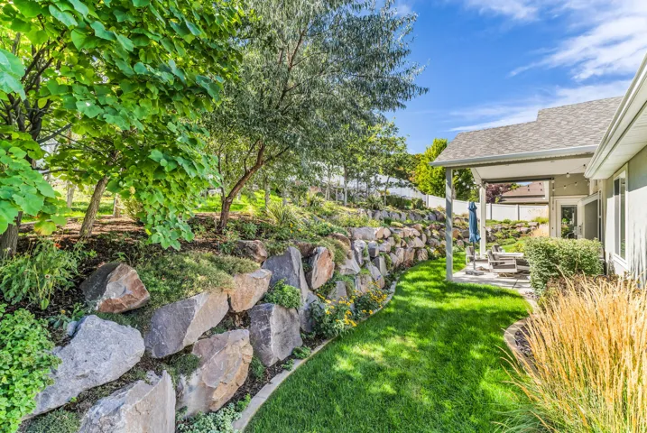 Terraced landscaping with a stacked stone retaining wall, mature trees, manicured lawn, and covered outdoor patio
