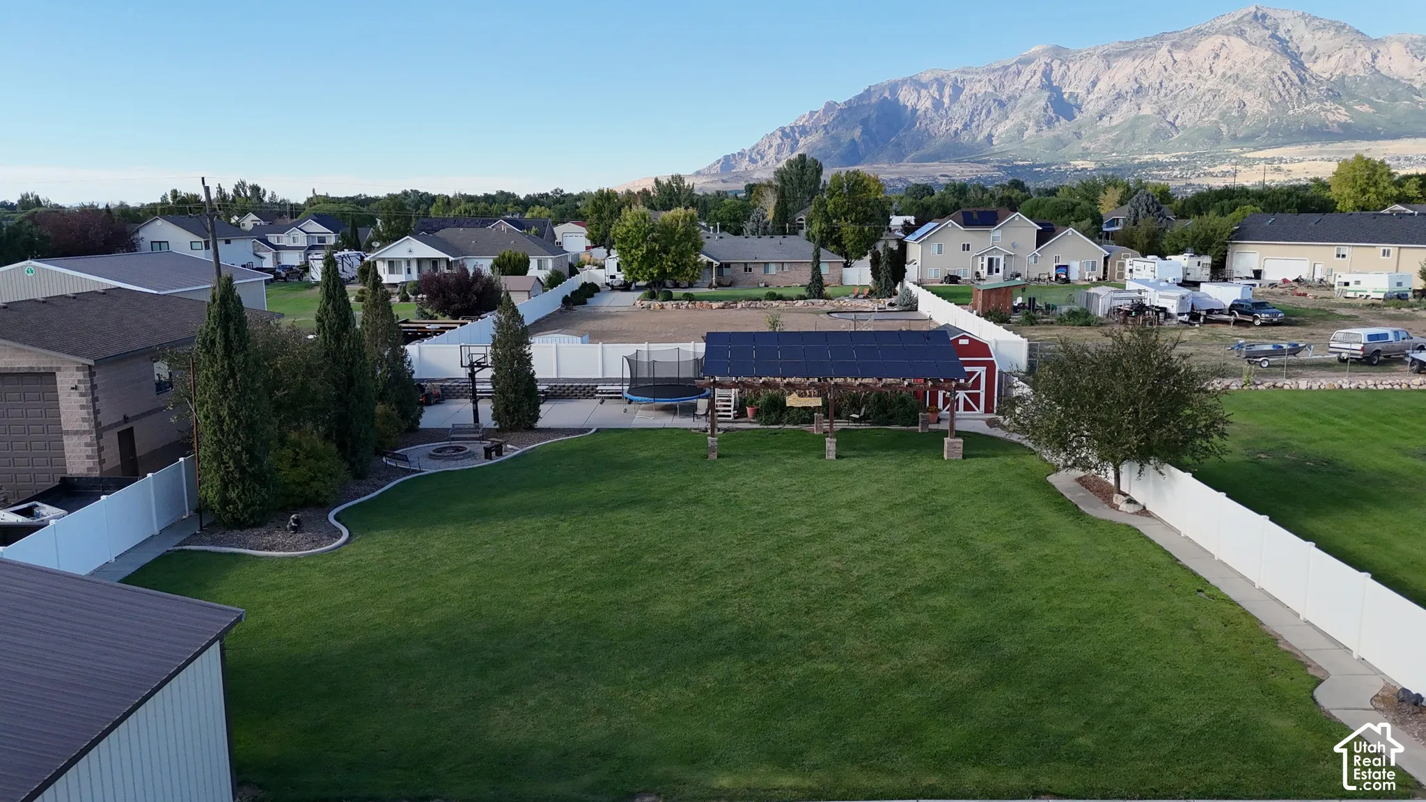 Expansive green lawn, white perimeter fencing, paved patio with fire pit, shaded pergola, and detached red shed