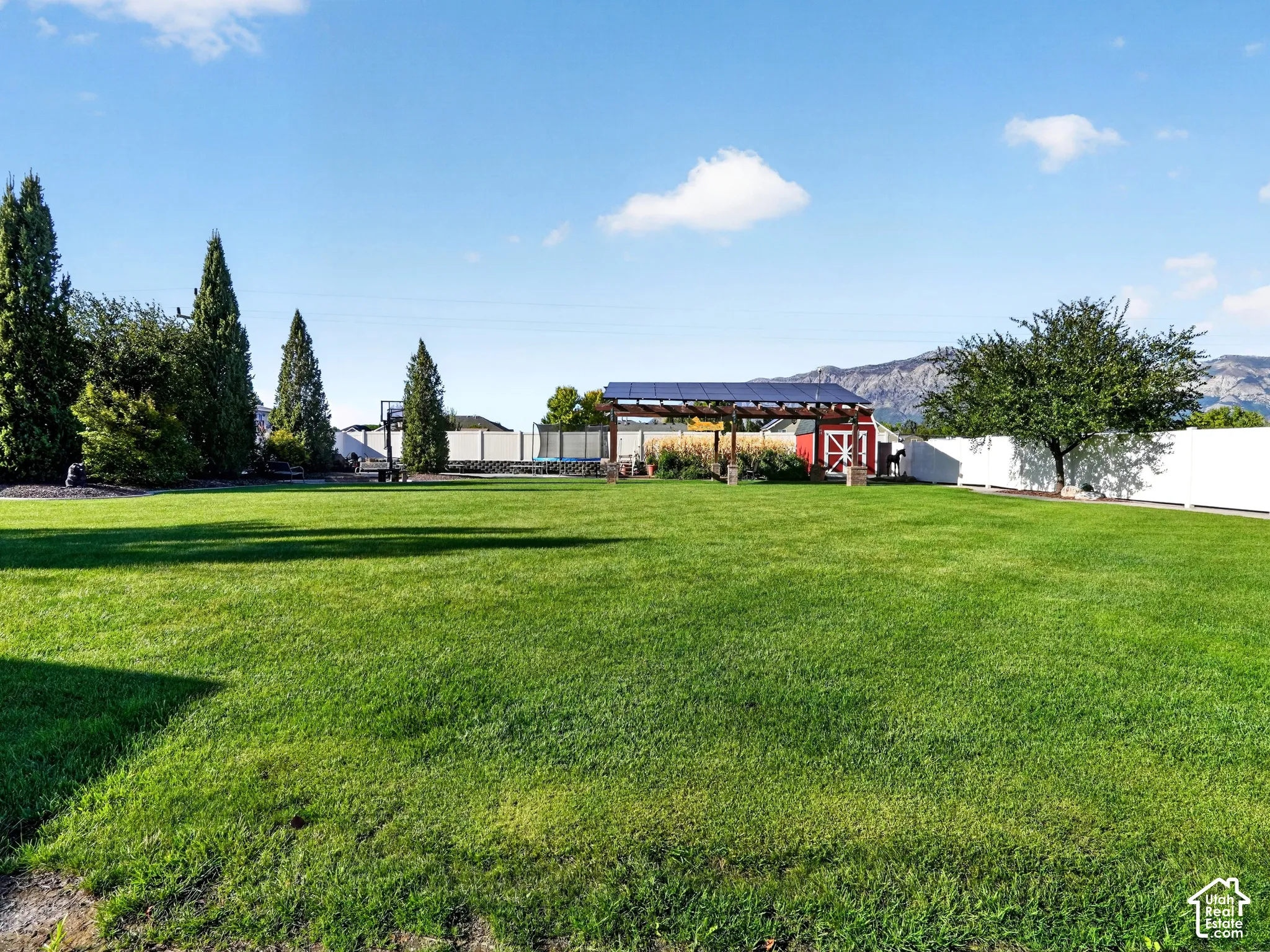 Expansive green lawn with mature evergreen trees, a white perimeter fence, a red barn structure, and a pergola with solar panels