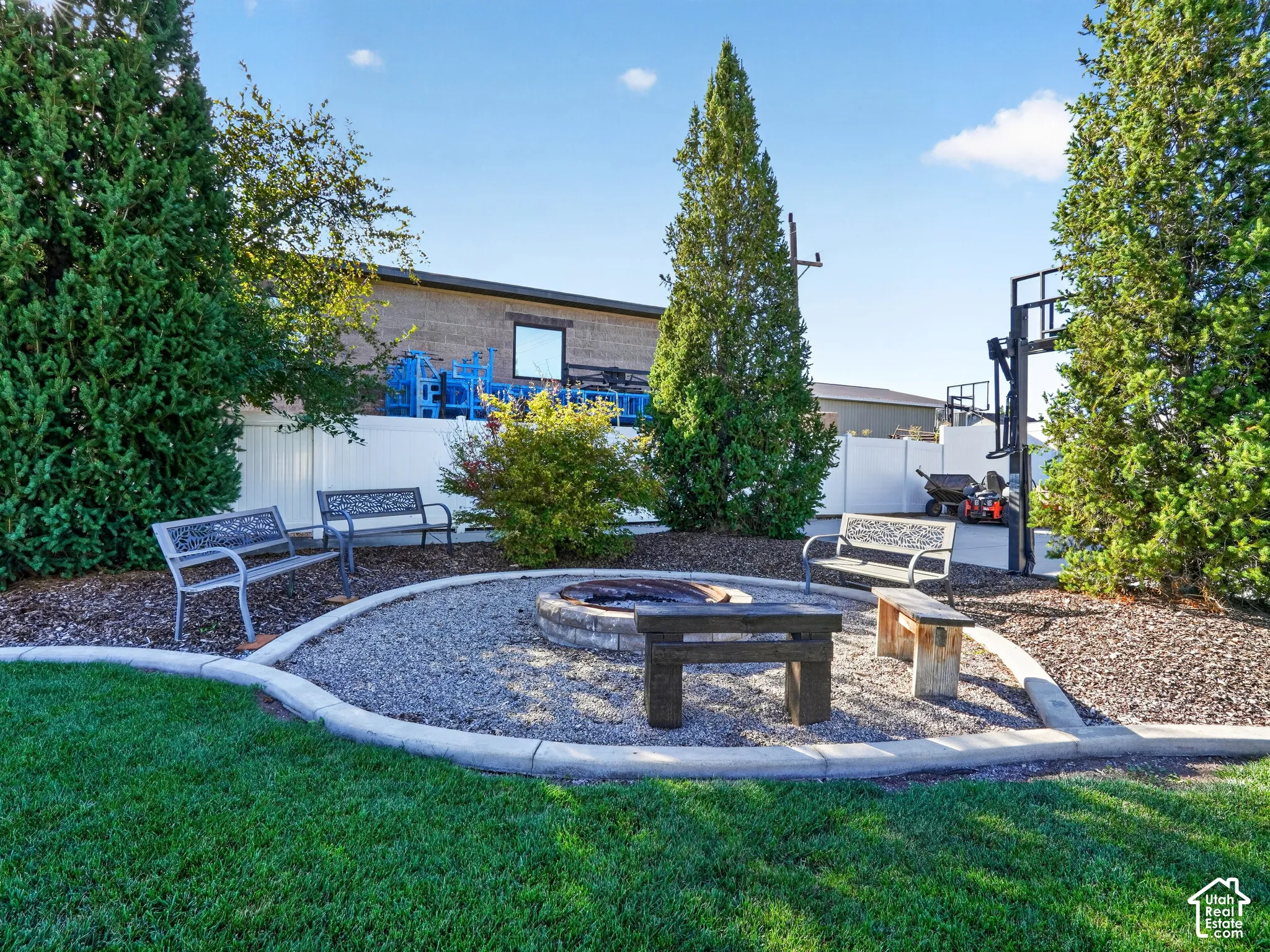 Manicured lawn with a gravel fire pit area, concrete curbing, mature evergreen trees, and a privacy fence