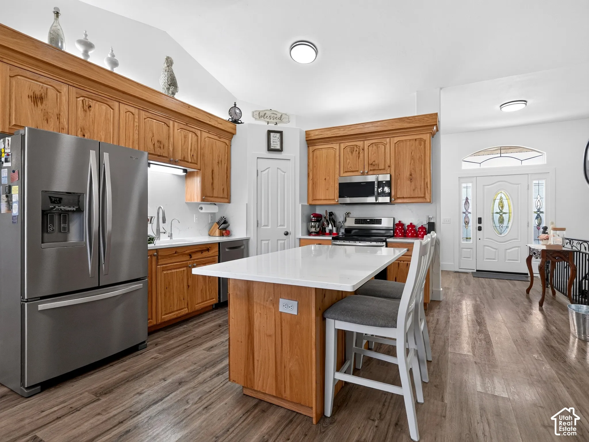 Wood-finish flooring throughout, kitchen island with seating, stainless steel appliances, wood cabinetry, and a decorative front door with stained glass accents