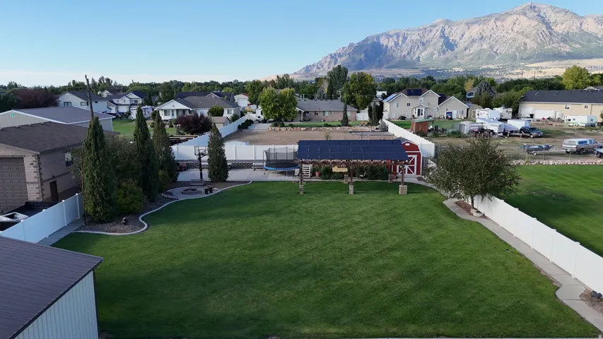 Expansive green lawn, white perimeter fencing, paved patio with fire pit, shaded pergola, and detached red shed