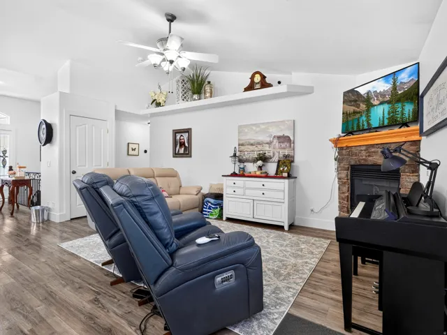 Living area featuring wood-finish flooring, a ceiling fan with integrated lighting, a stone-faced fireplace with a wooden mantel, and built-in floating shelves