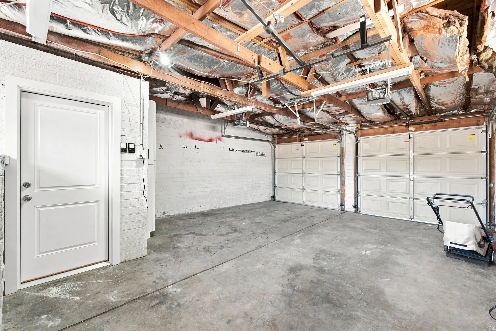 Two overhead garage doors, exposed wood beam ceiling, white painted brick walls, concrete flooring, and a white interior access door