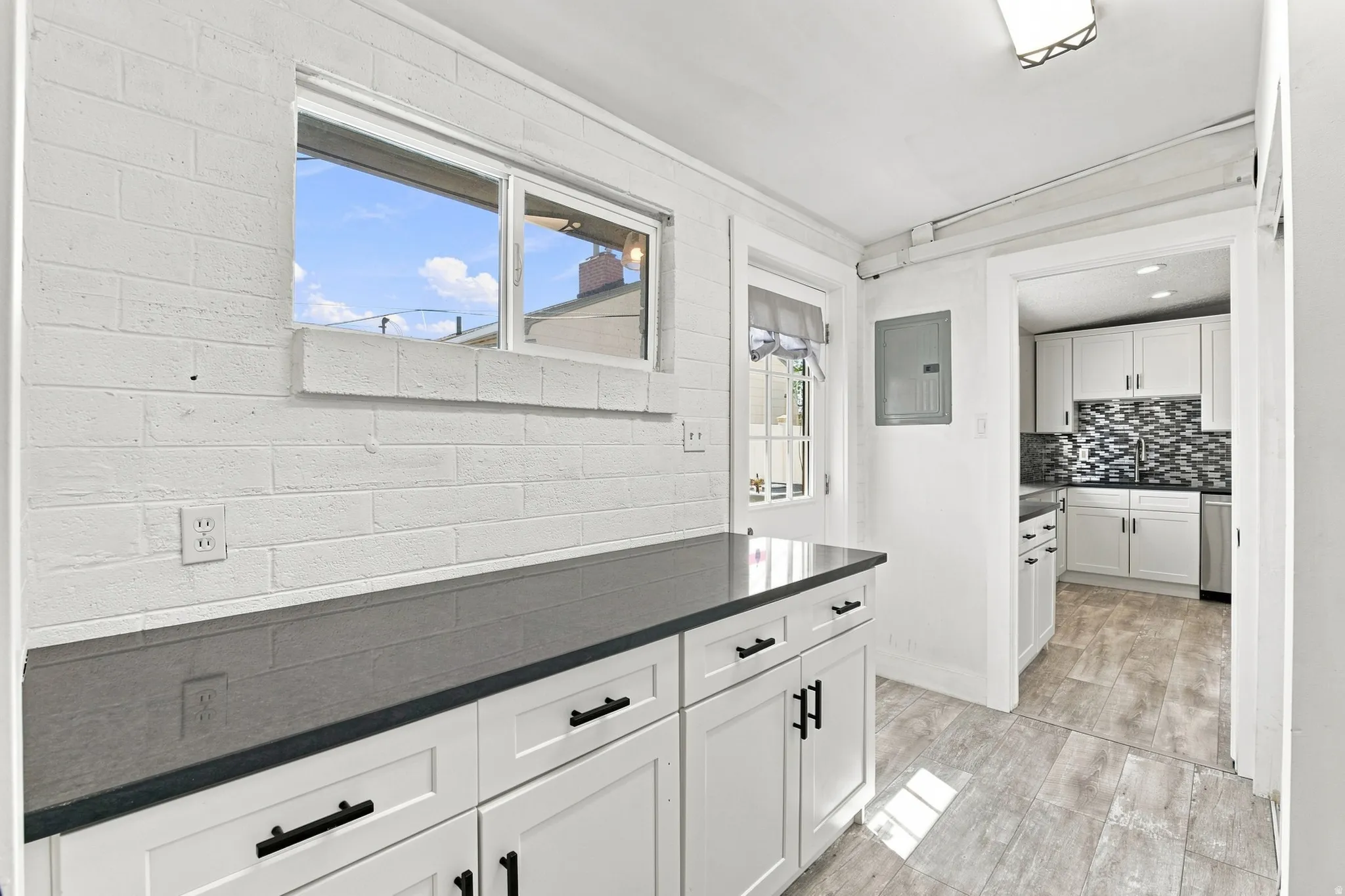 White cabinetry with black hardware, dark countertops, white painted brick wall, and wood-look flooring
