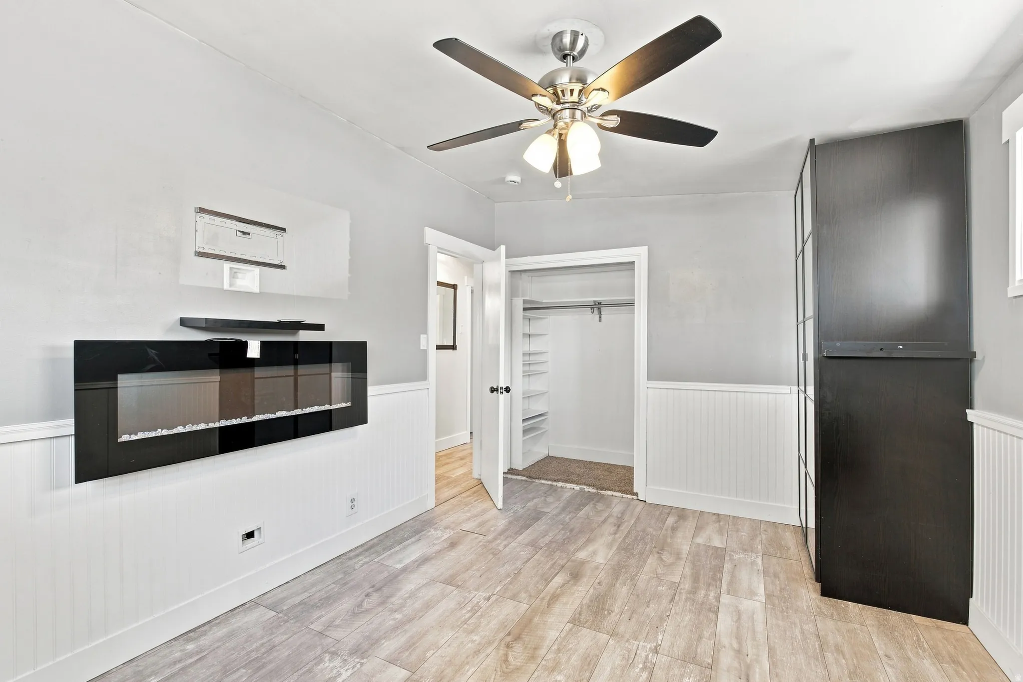 Wood-finish flooring, wainscoting, contemporary wall-mounted fireplace, ceiling fan with integrated lighting, and built-in closet shelving