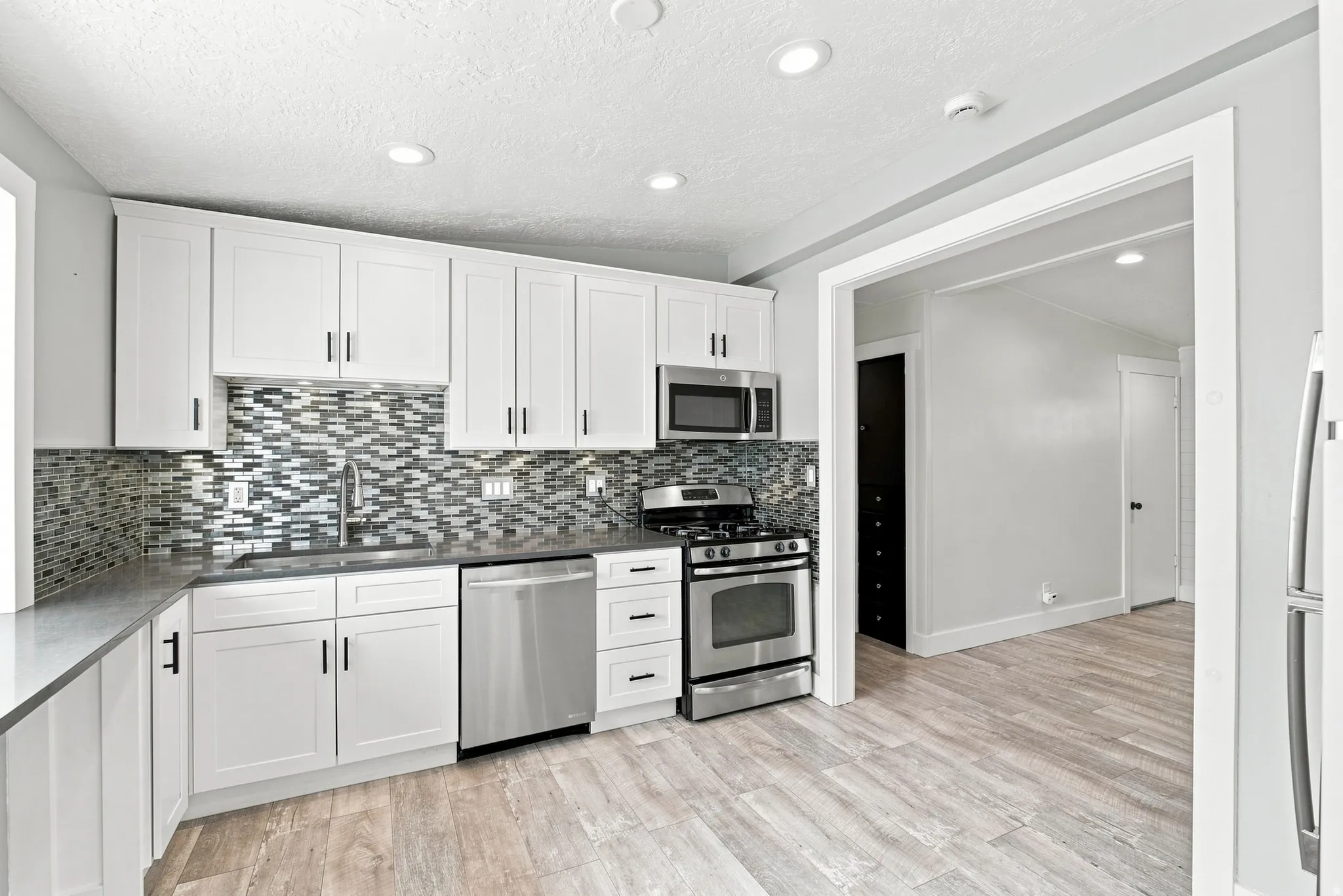 Modern kitchen with white cabinetry and dark hardware, stainless steel appliances, mosaic tile backsplash, and light wood-finish flooring