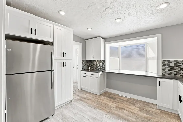 White shaker cabinetry with matte black hardware, stainless steel refrigerator, dark countertops, mosaic tile backsplash, and wood-finish flooring