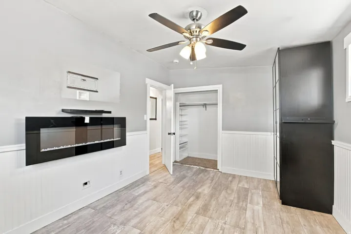 Wood-finish flooring, wainscoting, contemporary wall-mounted fireplace, ceiling fan with integrated lighting, and built-in closet shelving