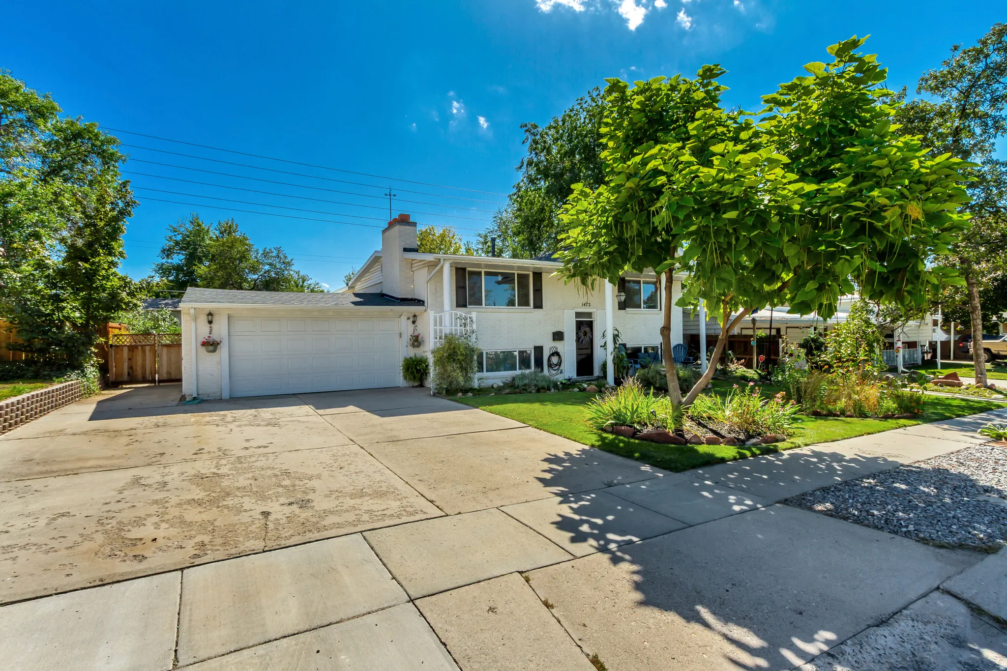 White exterior with a two-car garage, mature trees, landscaped front yard, and a concrete driveway
