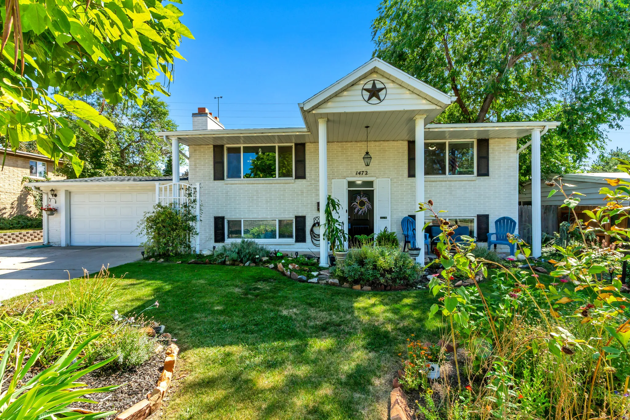 Split-level residence featuring a covered front porch, white brick exterior, attached garage, established landscaping, and mature trees