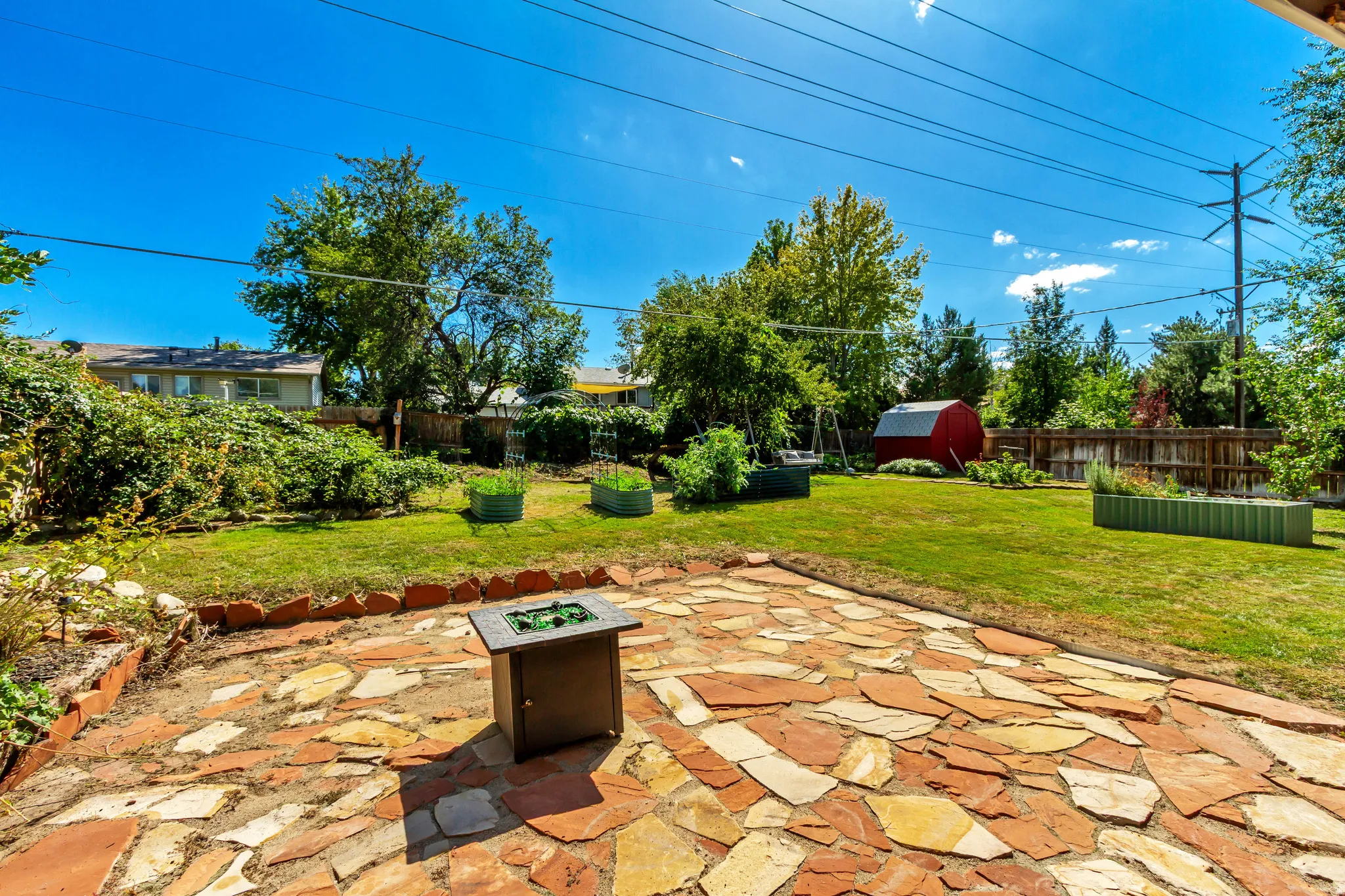 Expansive flagstone patio with a built-in fire pit, surrounded by a manicured lawn and raised garden beds
