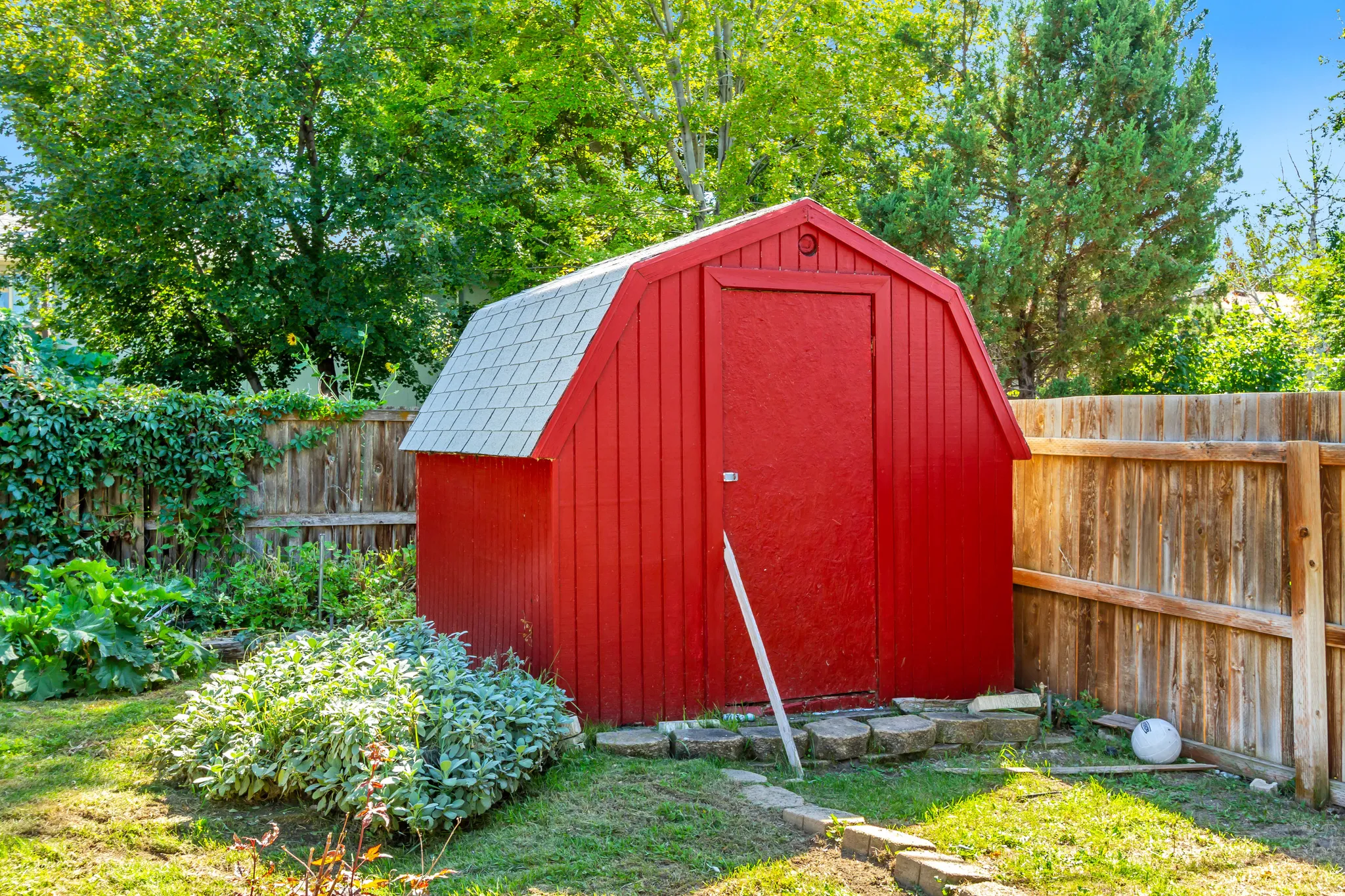 Red barn-style shed with a grey shingle roof, surrounded by a wooden privacy fence, mature trees, and garden plantings