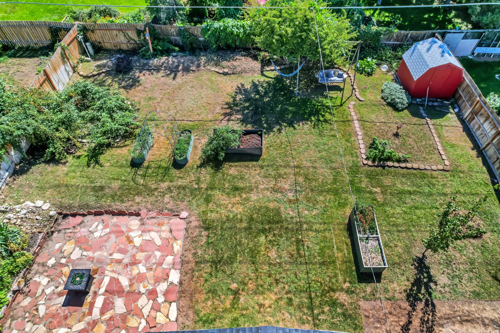 Expansive lawn featuring a flagstone patio, raised garden beds, mature trees, and a red storage shed