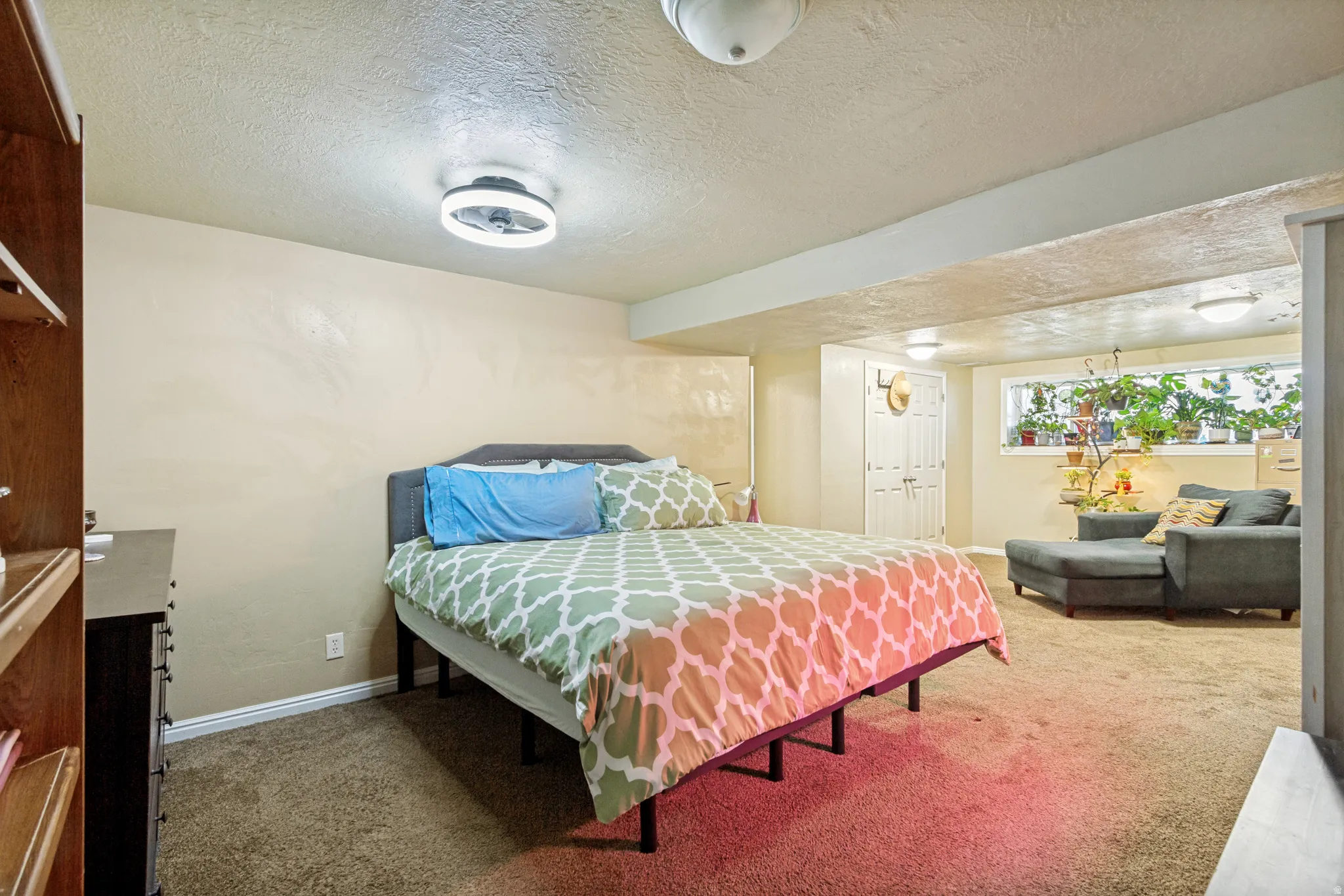 Carpeted room with a built-in wooden shelving unit, ceiling-mounted light fixture, and a window with natural light