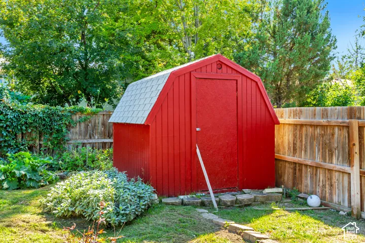 Red barn-style shed with a grey shingle roof, surrounded by a wooden privacy fence, mature trees, and garden plantings