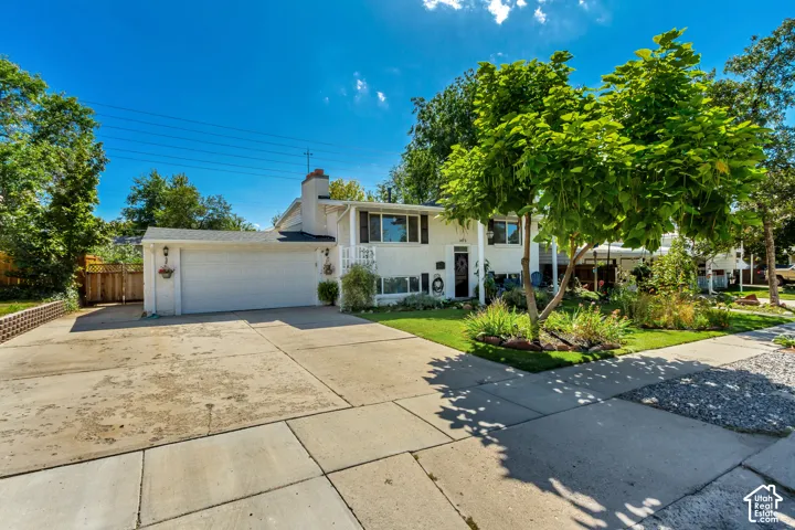 White exterior with a two-car garage, mature trees, landscaped front yard, and a concrete driveway